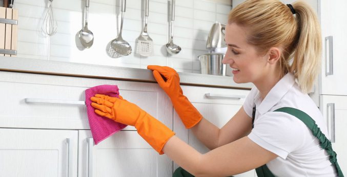 Young woman wearing orange latex gloves cleaning white shaker style cabinets.